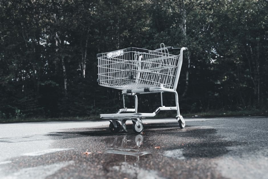 Lone shopping cart on wet road amidst forest, highlighting urban decay and loneliness.